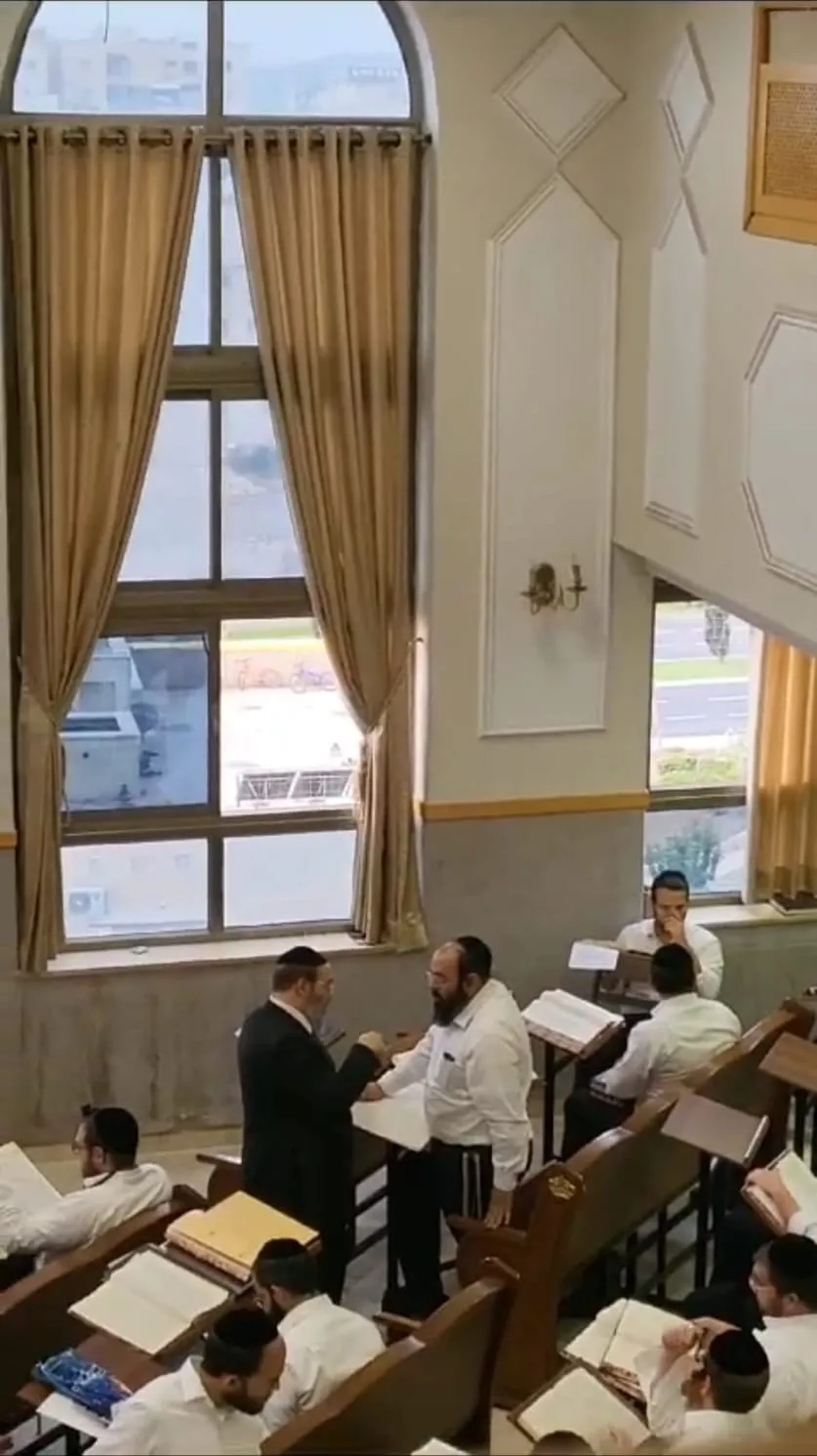 שני תלמידי חכמים עומדים בעיון על סטנדר, ומאחוריהם אברכים לומדים